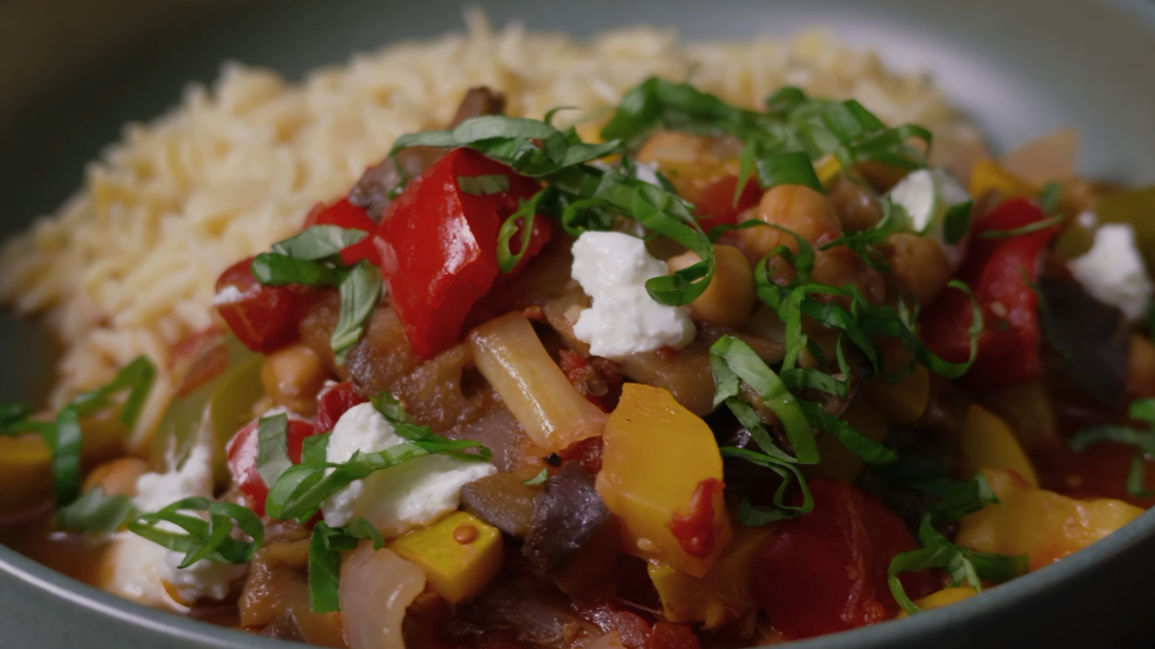 Bowl of ratatouille served over orzo with chickpeas, red and yellow peppers, eggplant, fresh basil and crumbled goat cheese